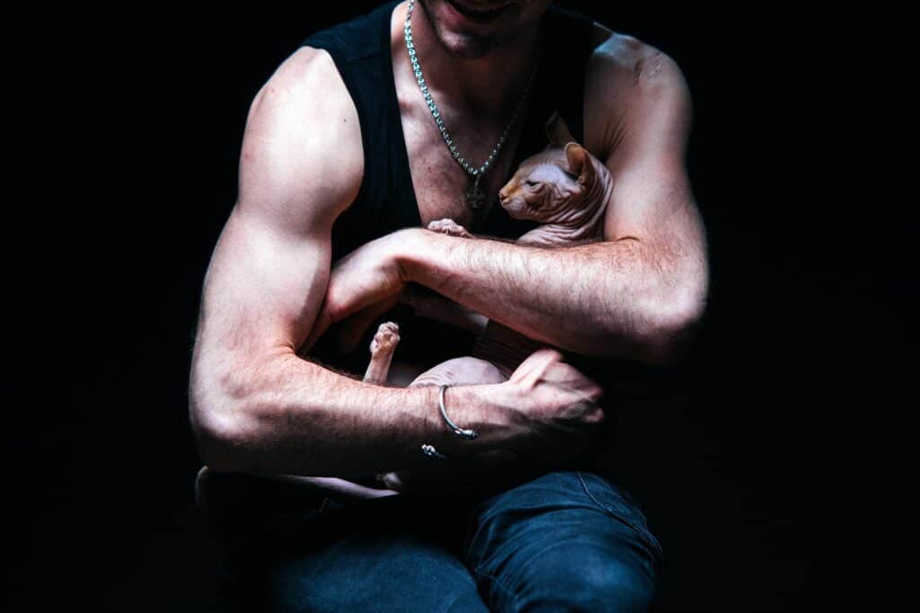 man in a black sleeveless shirt cuddling a sphynx cat, black background